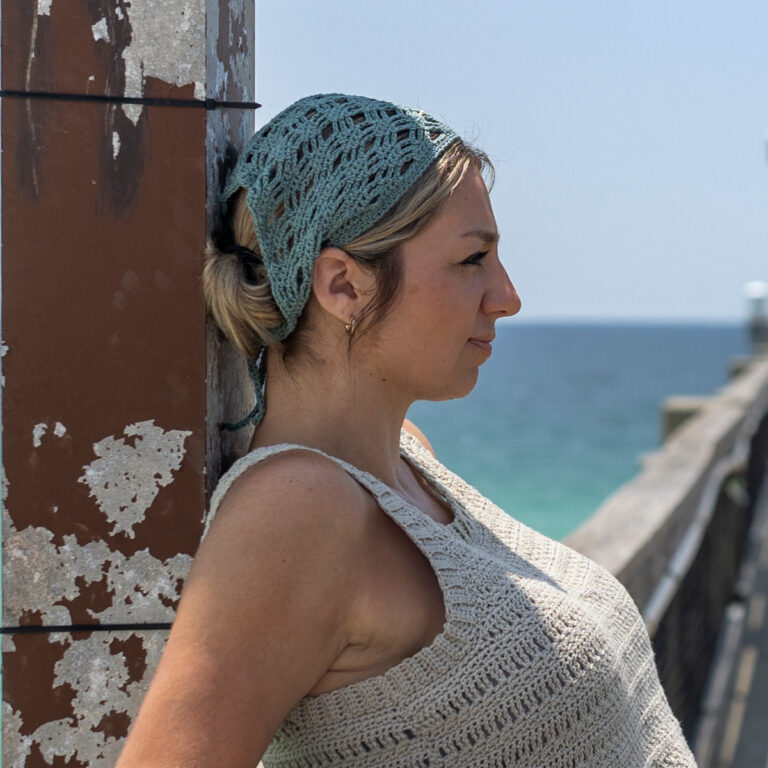 Girl leaning against a bridge wearing a grey crochet top with her hair tied back under a blue crochet bandana.