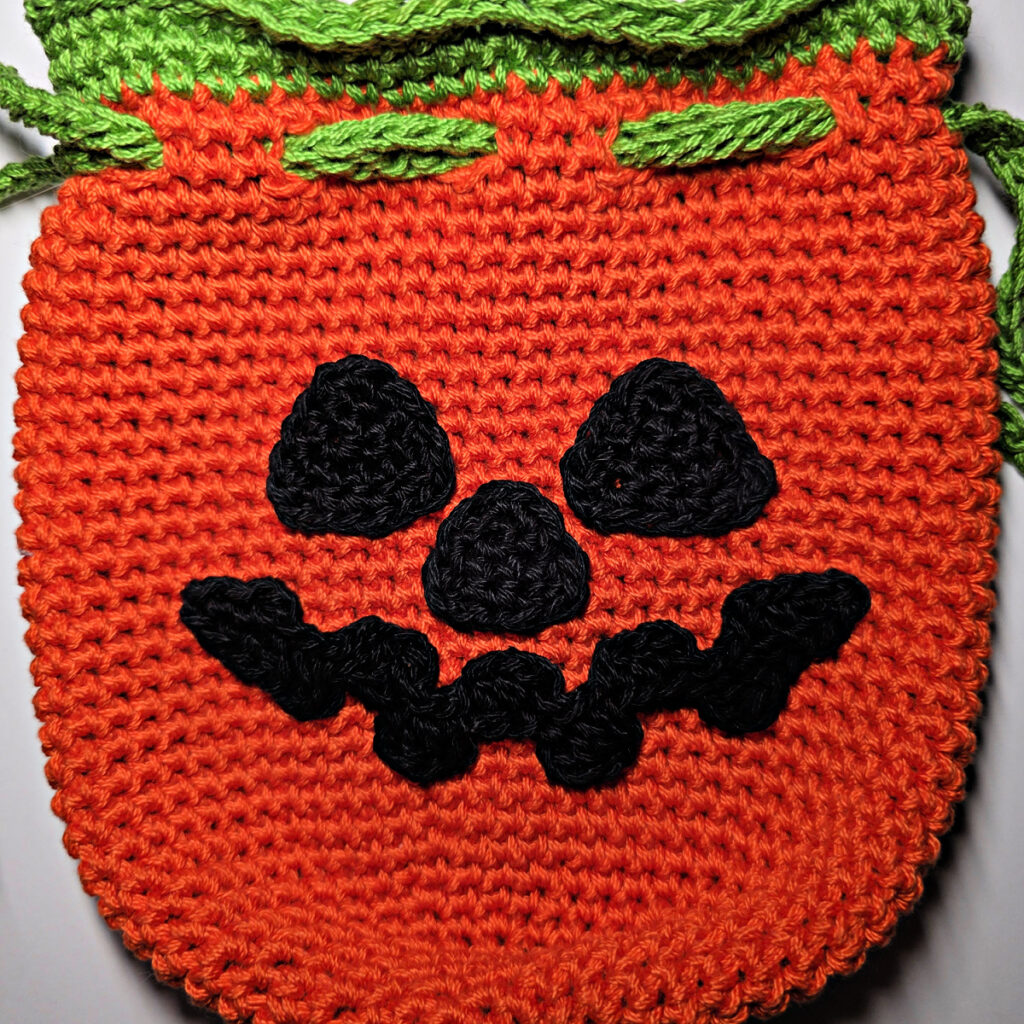 A jack-o-lantern crochet treat bag with a green drawstring.