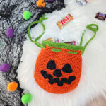 A crochet Jack-O-Lantern pumpkin treat bag full of candy against a white faux fur background.