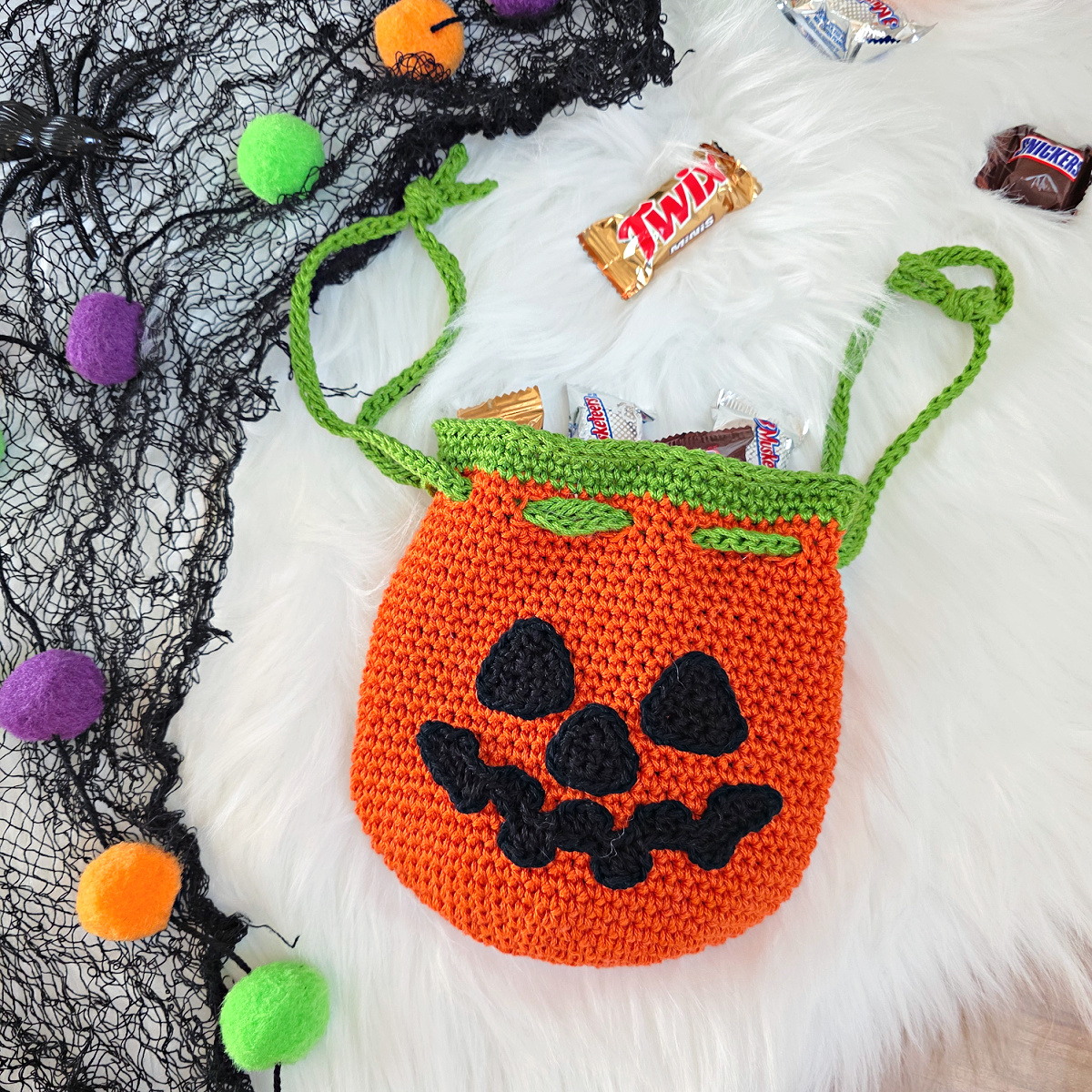 A crochet Jack-O-Lantern pumpkin treat bag full of candy against a white faux fur background.
