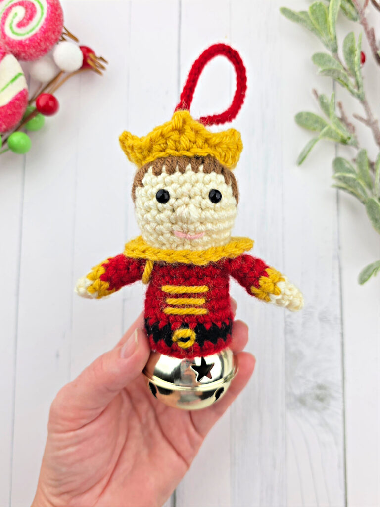 A hand holding a Nutcracker Prince crochet jingle bell ornament near small Christmas decorations.