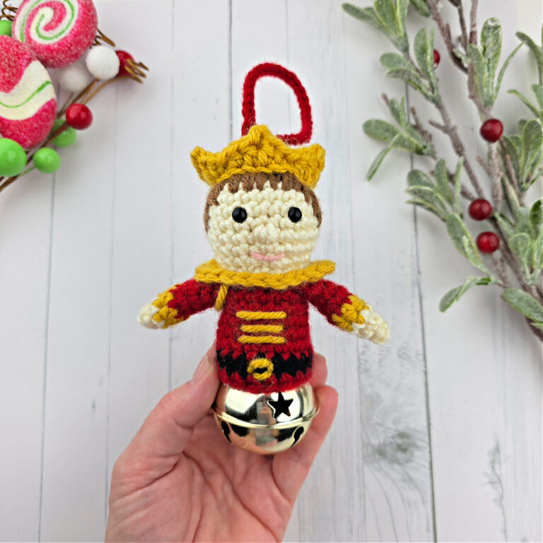 A hand holding a Nutcracker Prince crochet jingle bell ornament near small Christmas branches.