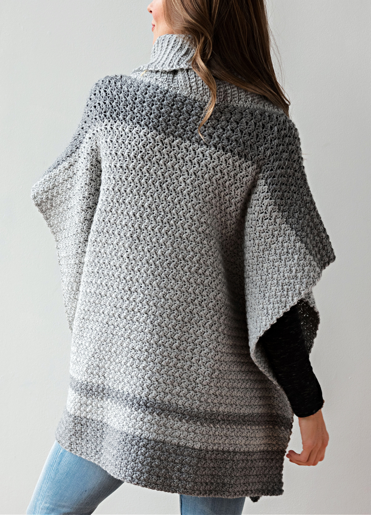 Woman wearing a two toned crochet poncho with a cowl neck and facing away from the camera.