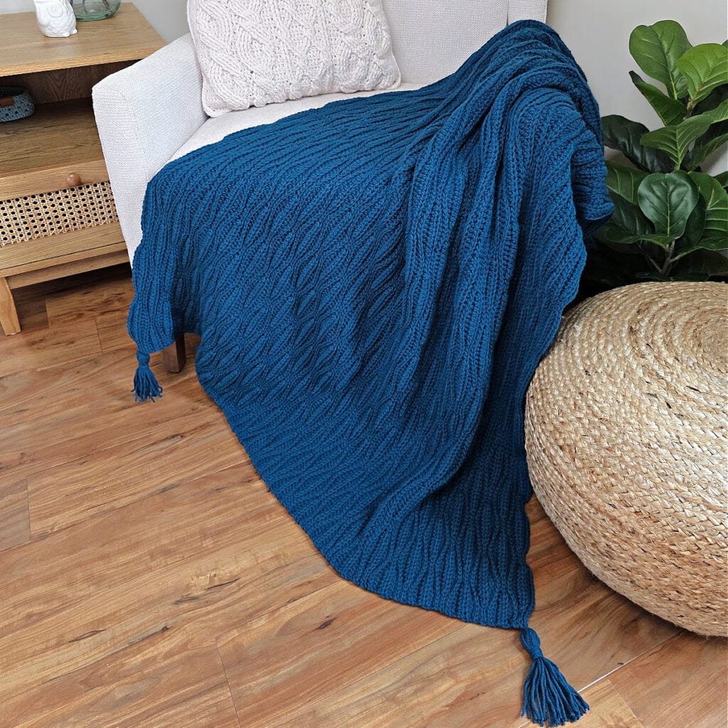 A dark blue crochet blanket draped over a sofa chair and wooden floor with a cushion next to it.