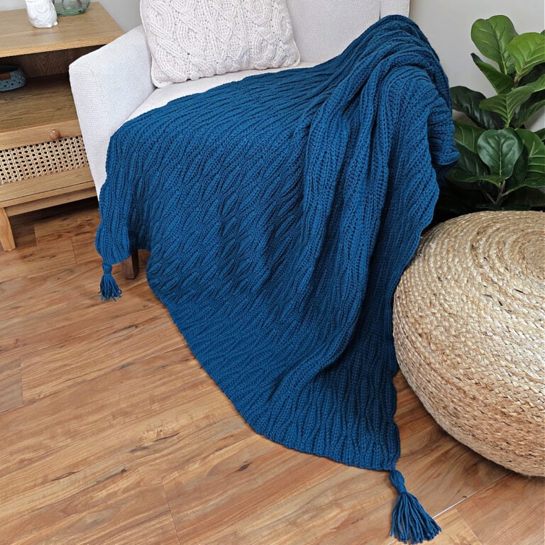 A dark blue crochet blanket draped over a sofa chair and wooden floor with a cushion next to it.
