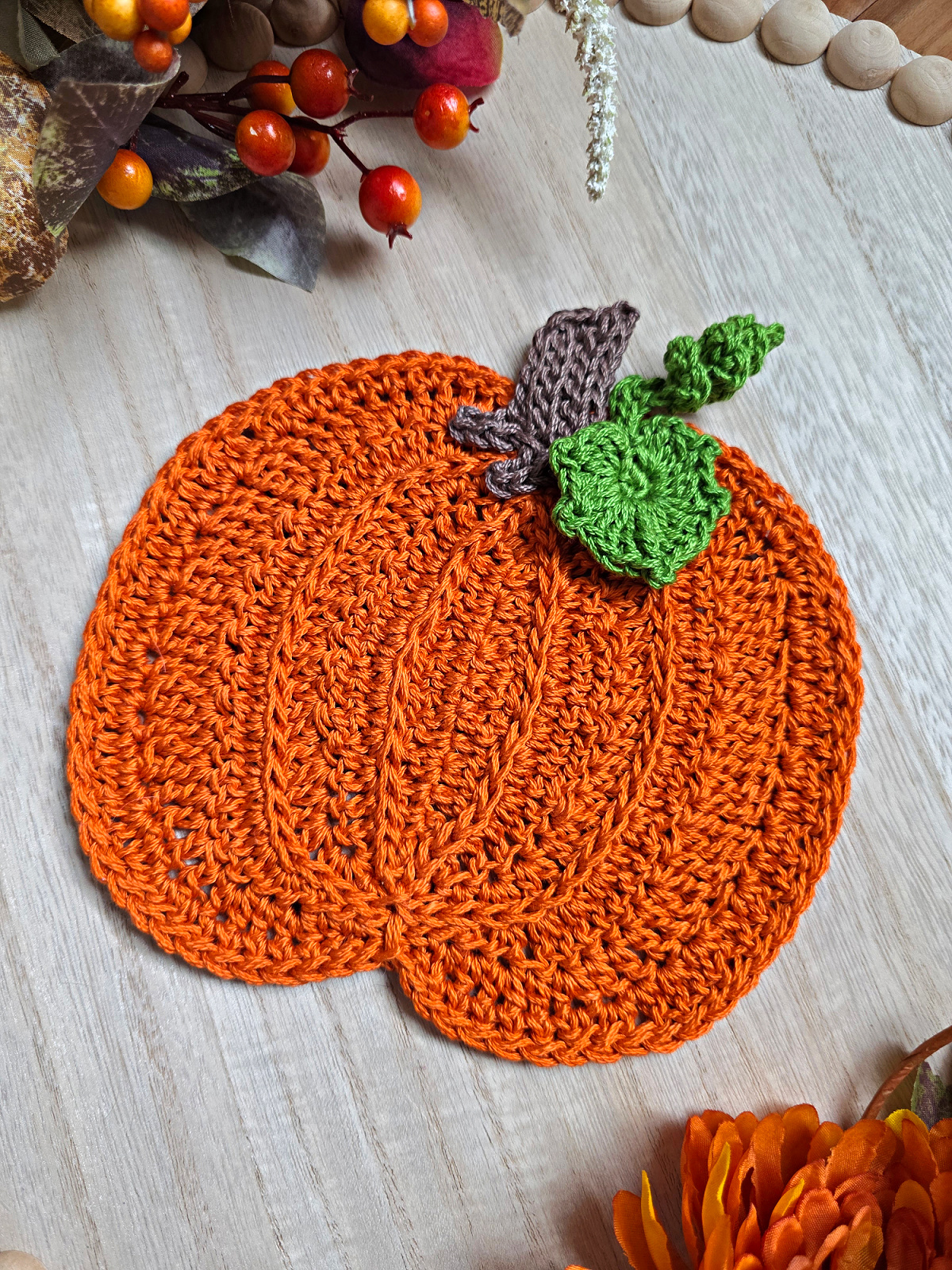 Large orange crochet pumpkin with a brown stem and green leaves against a wooden background with fall decor around it.