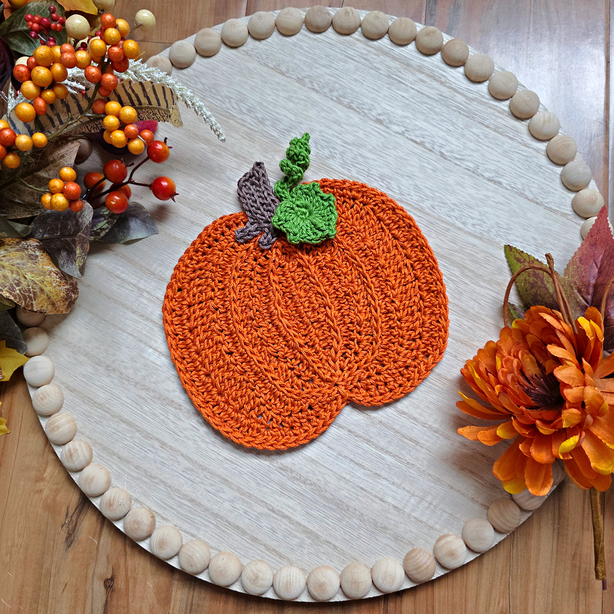Large orange crochet pumpkin with a brown stem and green leaves against a white wooden tray with fall decor around it.