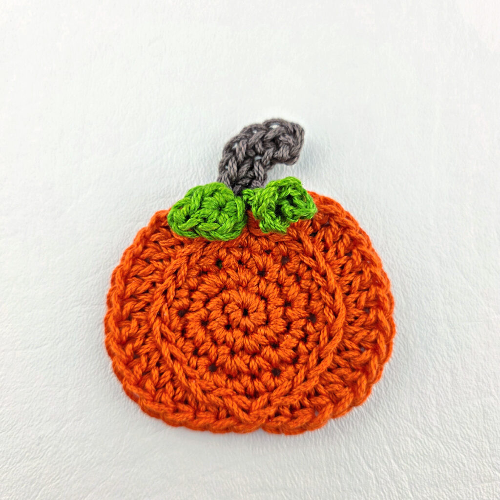 A crochet pumpkin applique complete with a pumpkin leaf and tendril.