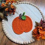 Large orange crochet pumpkin with a brown stem and green leaves against a white wooden tray with fall decor around it.