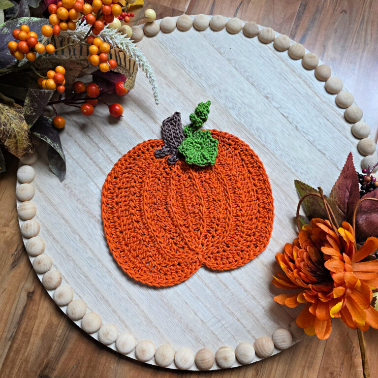Large orange crochet pumpkin with a brown stem and green leaves against a white wooden tray with fall decor around it.