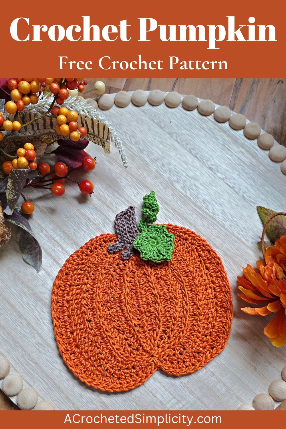 Large orange crochet pumpkin with a brown stem and green leaves against a white wooden tray with fall decor around it.