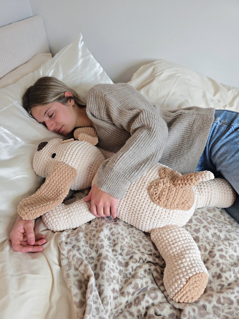Girl holding a plush brown and white crochet dog while laying on a bed with white and leopard print sheets.