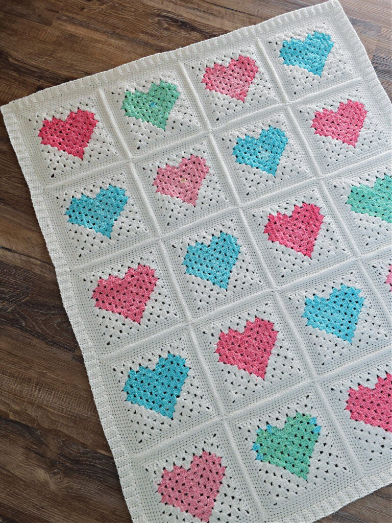 White crochet quilt made from pink and blue heart motifs spread out on a wooden floor.