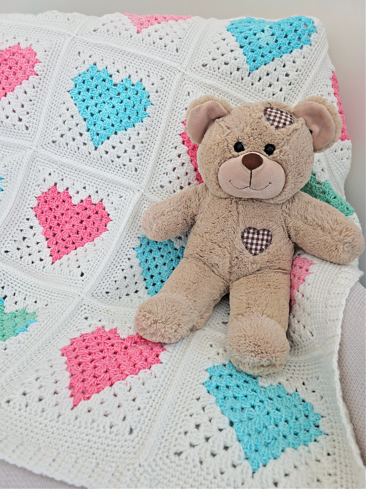 White crochet quilt made from pink and blue heart motifs spread on a sofa with a brown teddy bear on top.