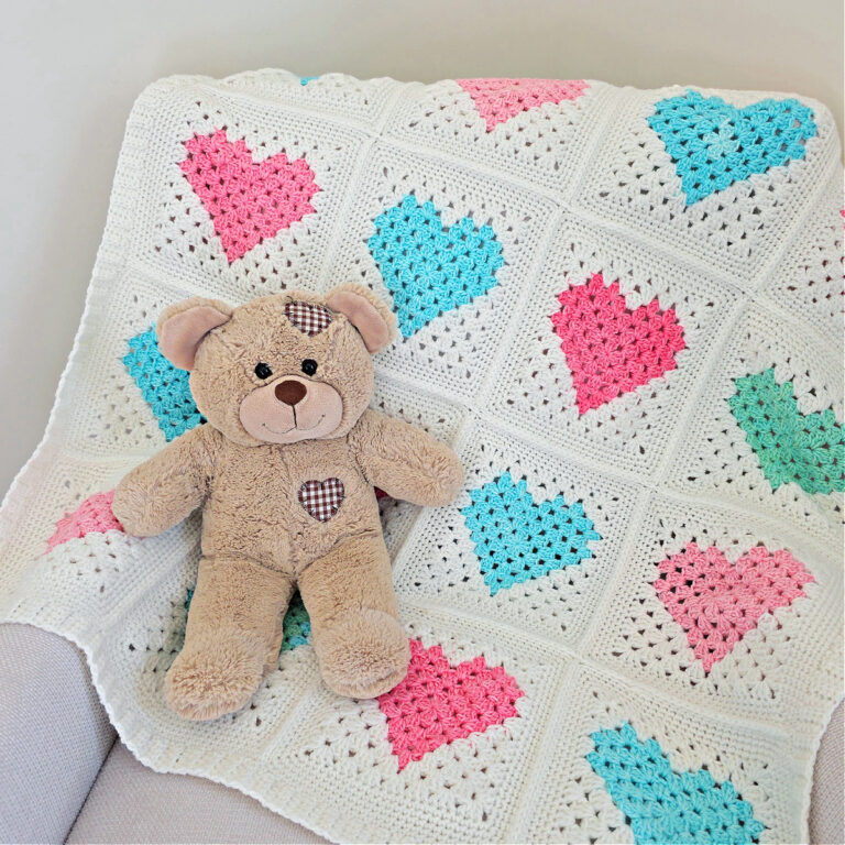 White crochet quilt made from pink and blue heart motifs spread on a sofa with a brown teddy bear on top.