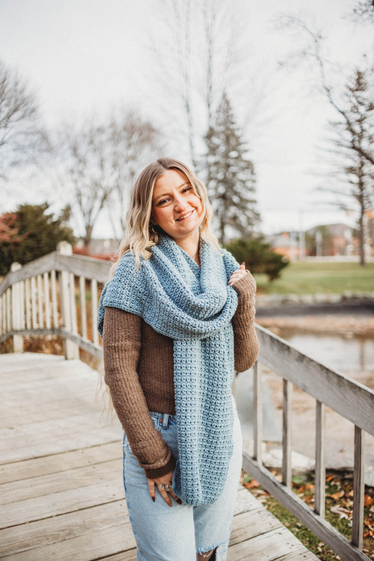 Woman wearing a brown sweater and blue jeans with a large blue crochet scarf wrapped around her neck standing on a wooden bridge.