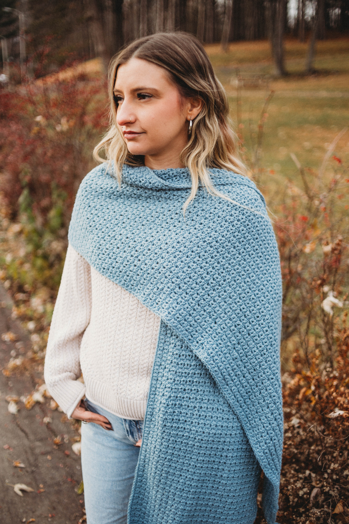 Woman wearing a white sweater with a large blue crochet scarf wrapped around her neck, turned to the side from the camera.