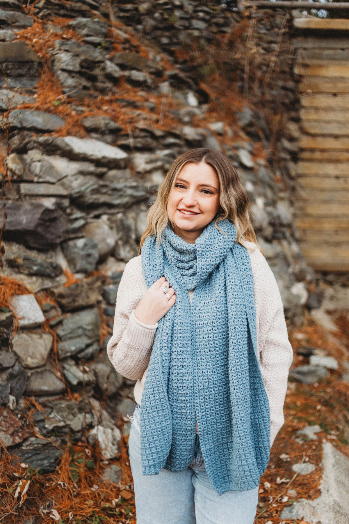 Woman wearing a white sweater and blue jeans with a large blue crochet scarf wrapped around her neck.