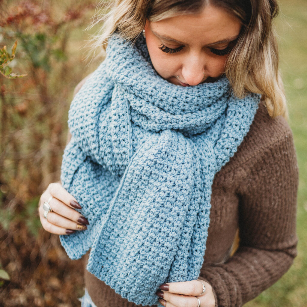 Woman wearing a brown sweater with a large blue crochet scarf wrapped around her neck, holding the edges, standing in a yard.