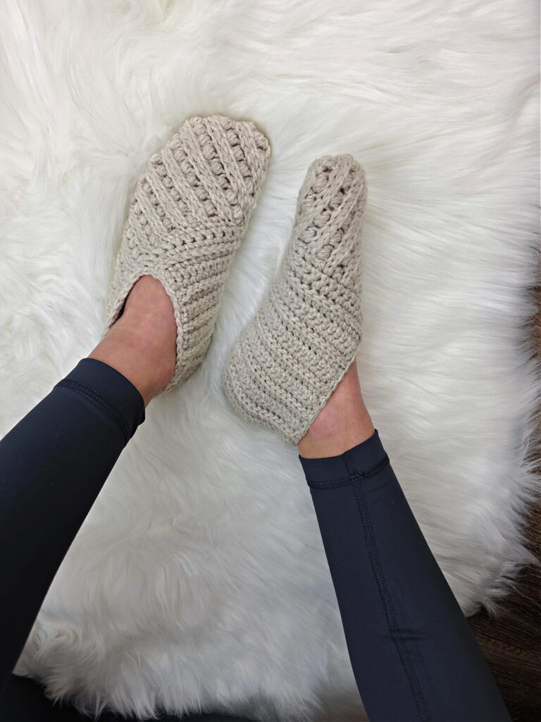 Model wearing grey crochet slippers on feet against a white plush background.