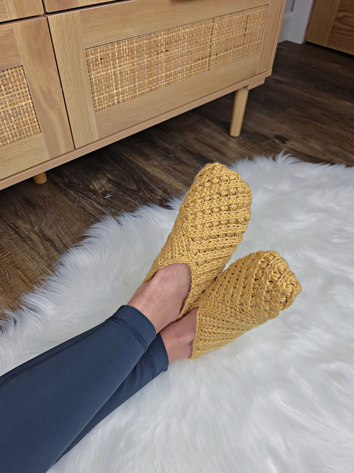 Model wearing mustard crochet slippers on feet against a white plush background.