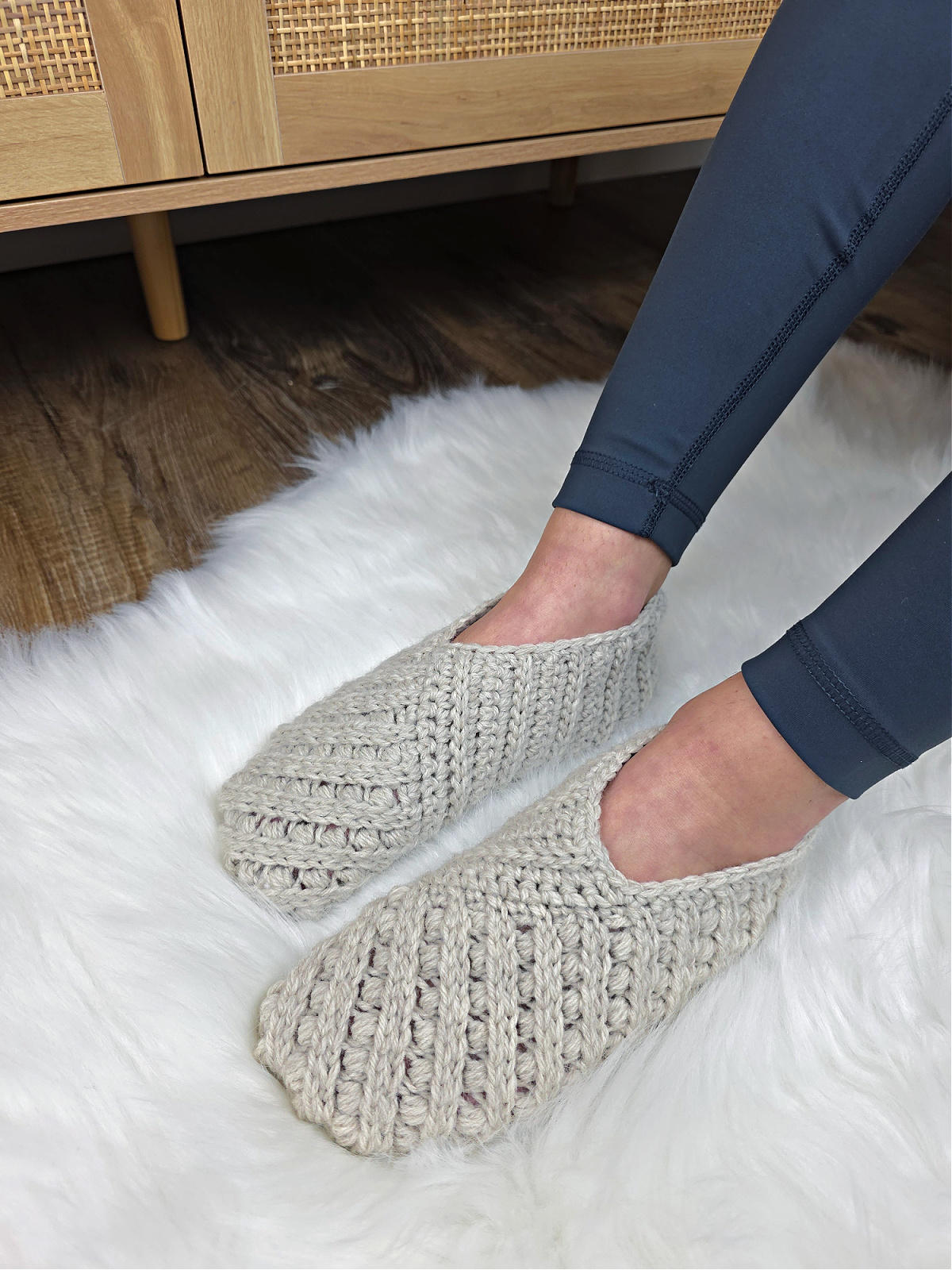 Model wearing grey crochet slippers on feet with a wooden cabinet in the background.