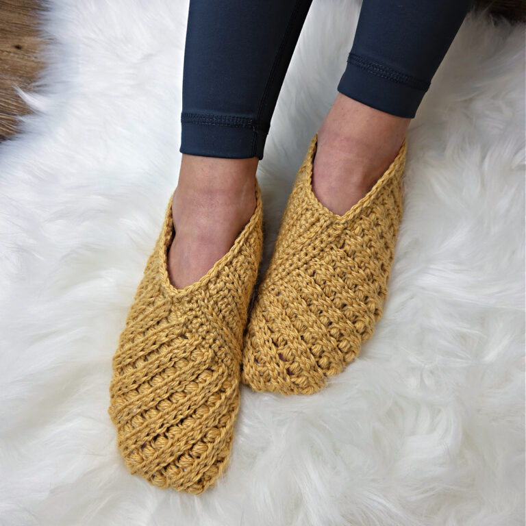 Model wearing mustard crochet slippers on feet against a white plush background.