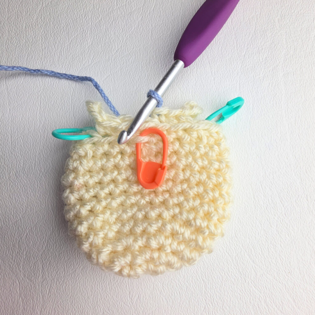 Small ivory crochet head for an ornament and a purple crochet hook and stitch markers joining blue yarn.