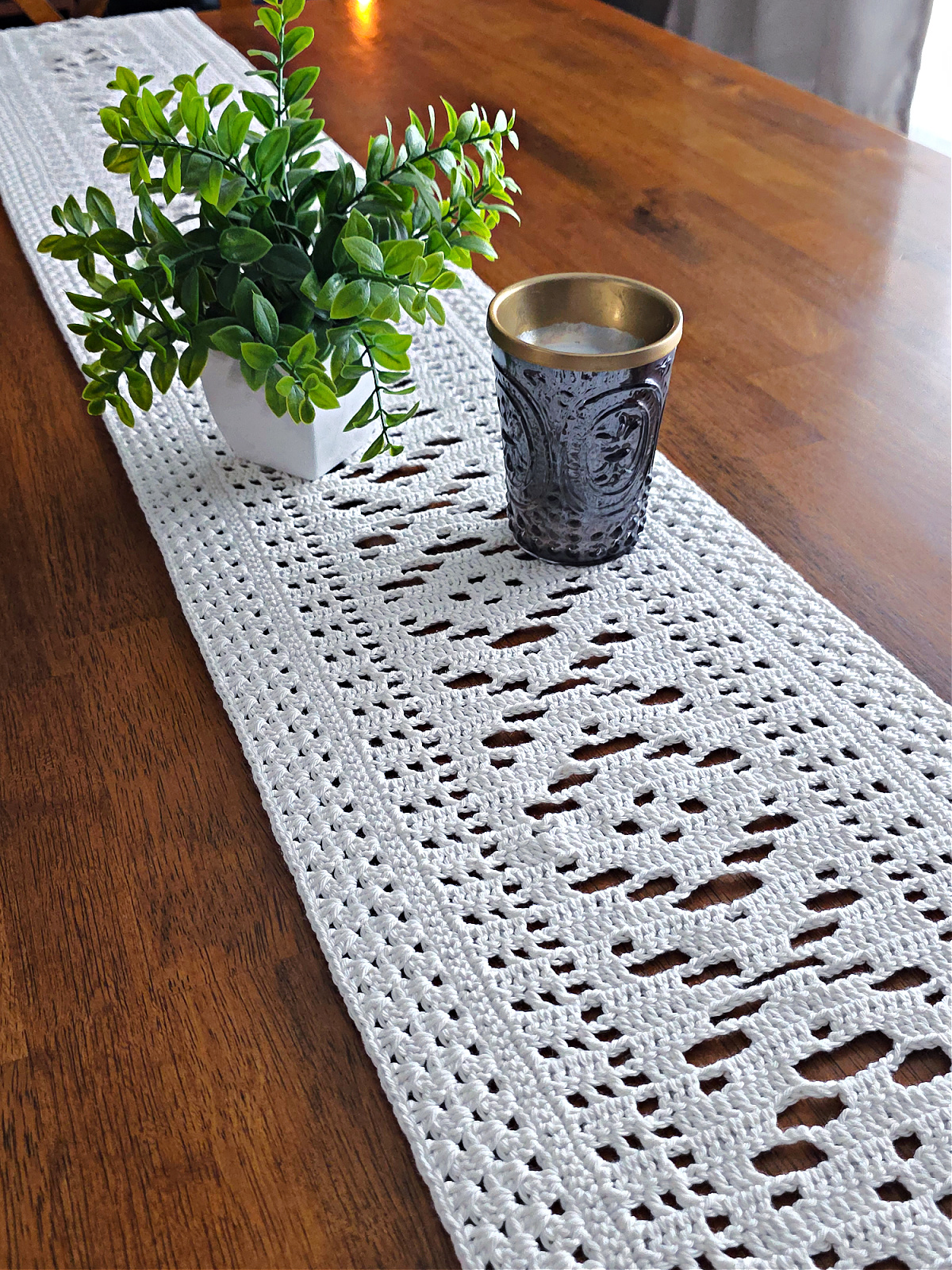 White lacy crochet table runner on a wooden table with a vase and silver candle on top.