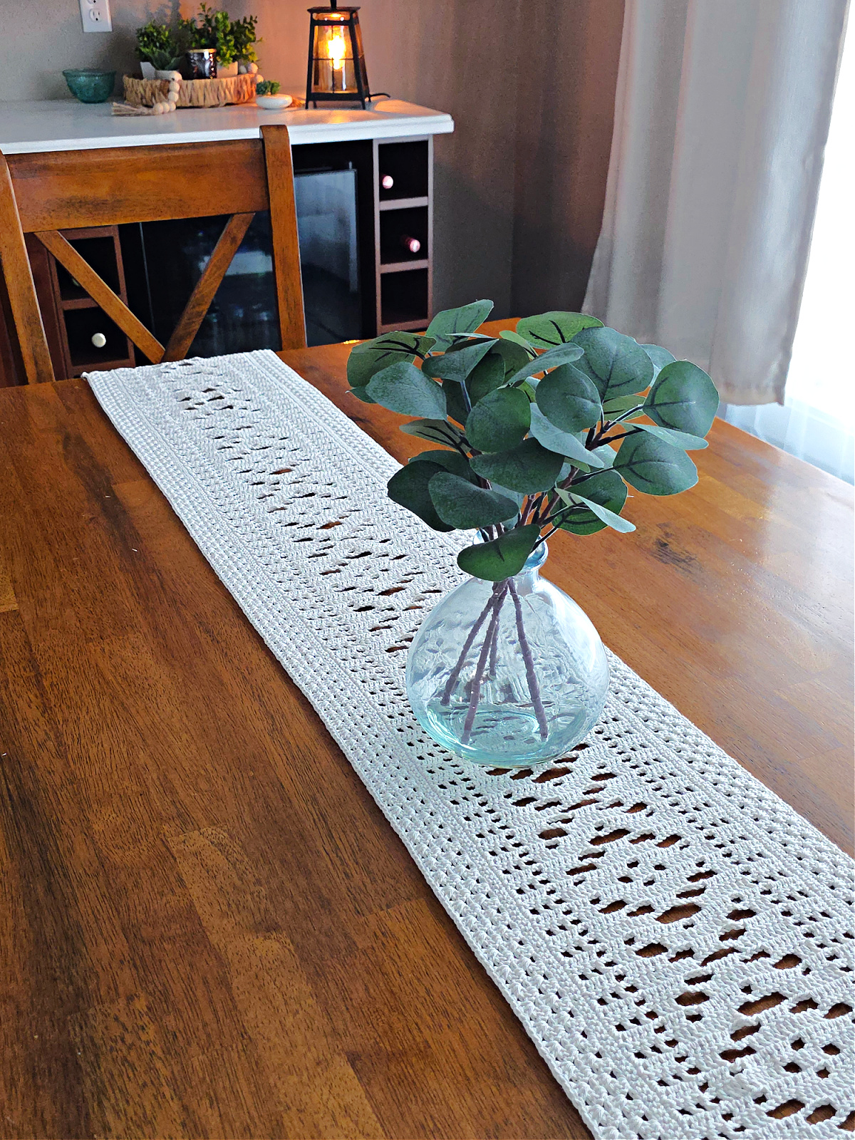 White lacy crochet table runner on a wooden table with a vase on top.