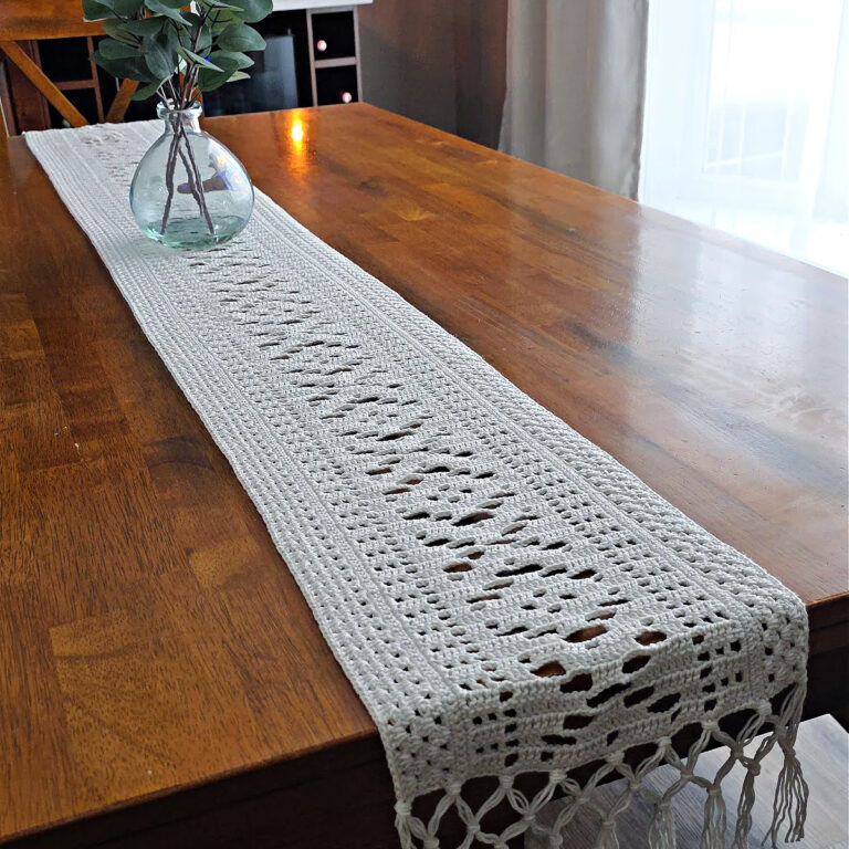 White lacy crochet table runner on a wooden table with a vase on top.