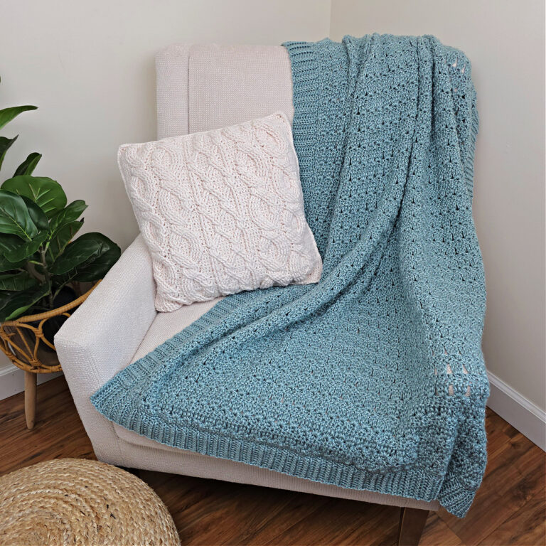 Blue crochet throw blanket with a white cushion on a white sofa with a plant next to it.