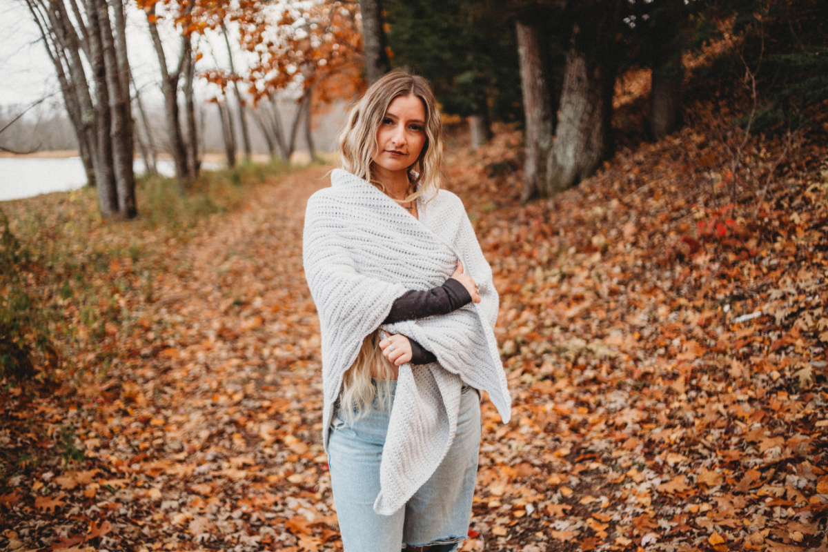 A woman standing on a path of fallen leaves wearing a grey crochet triangular shawl.