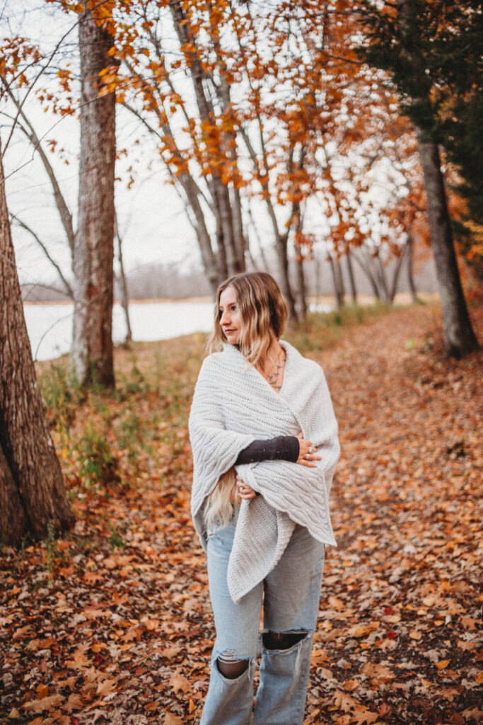 A woman standing on a path of fallen leaves wearing a grey crochet triangular shawl.
