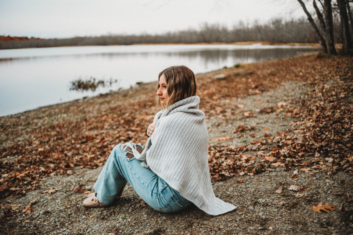 A woman sitting on the ground beside a lake wearing a grey crochet triangular shawl and jeans.