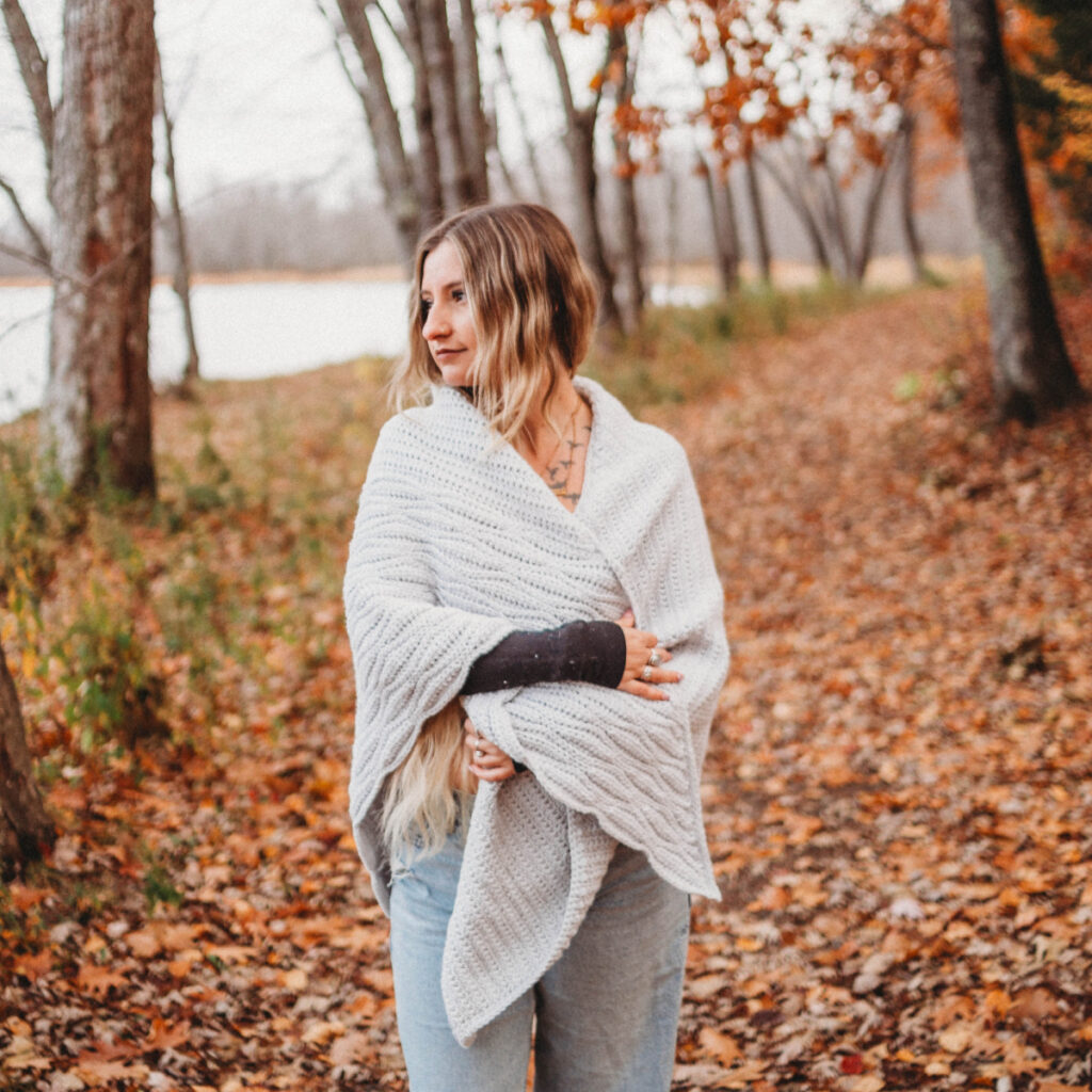A woman standing on a path of fallen leaves wearing a grey crochet triangular shawl.