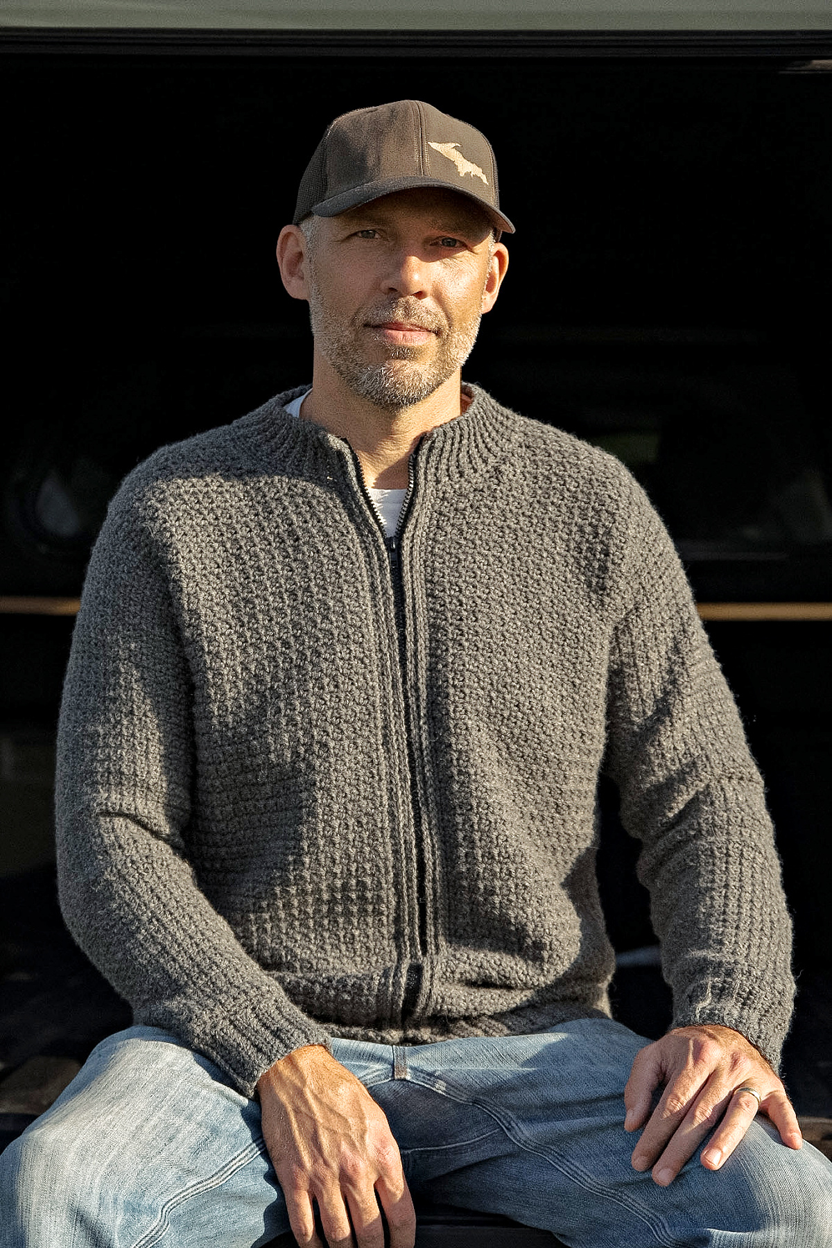 Man sitting and wearing a grey colored crochet mens cardigan with a grey hat and blue jeans.