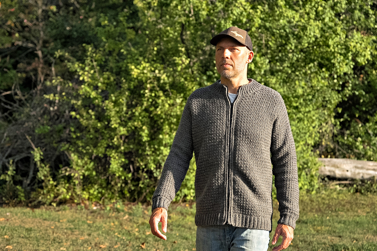 Man walking in the forest wearing a grey colored crochet mens cardigan with a grey hat and blue jeans.