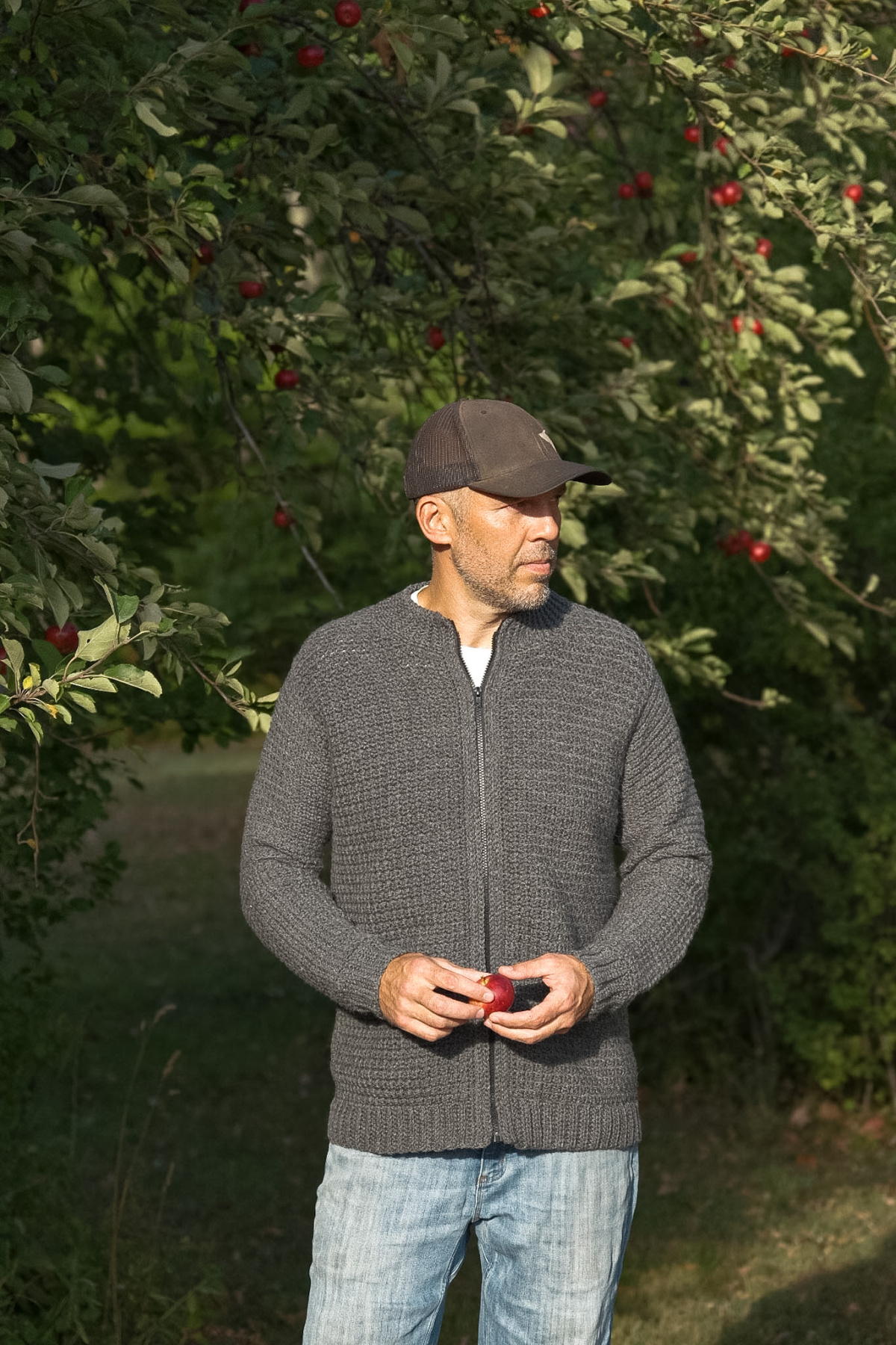 Man in the forest wearing a grey colored crochet mens cardigan with a grey hat and blue jeans.