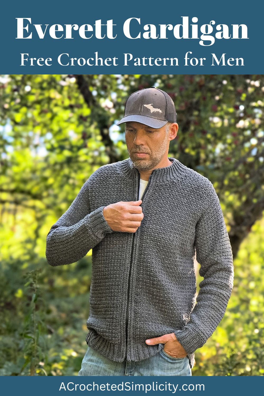 Man in the forest wearing a grey colored crochet mens cardigan, holding the zipper of the cardigan, with a grey hat and blue jeans.