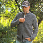 Man in the forest wearing a grey colored crochet mens cardigan, holding the zipper of the cardigan, with a grey hat and blue jeans.