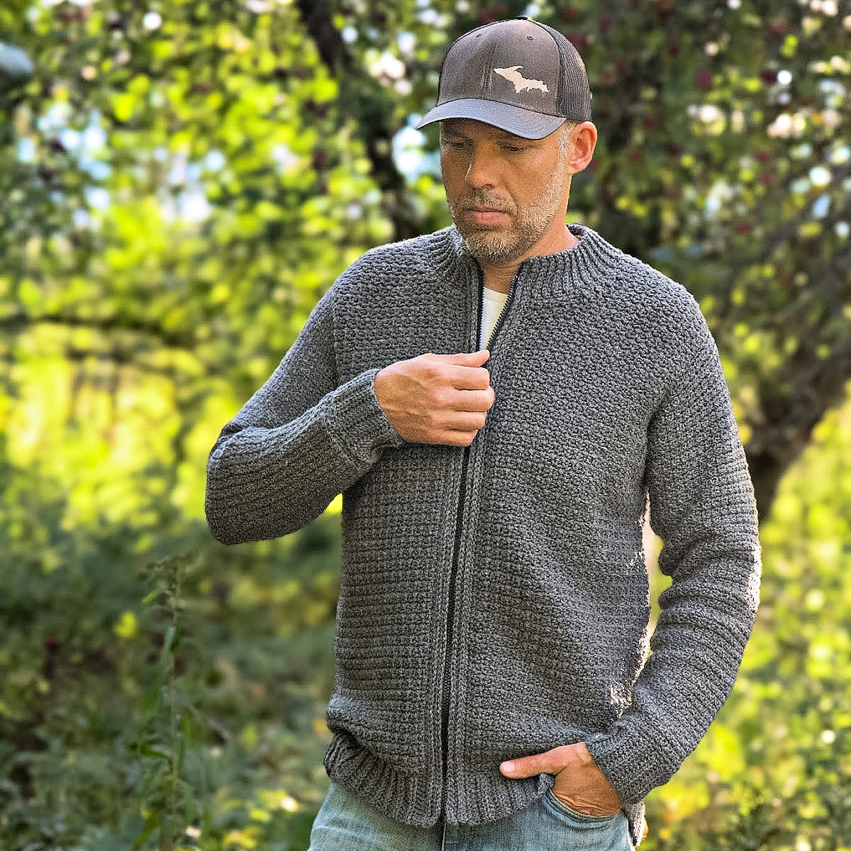 Man in the forest wearing a grey colored crochet mens cardigan, holding the zipper of the cardigan, with a grey hat and blue jeans.