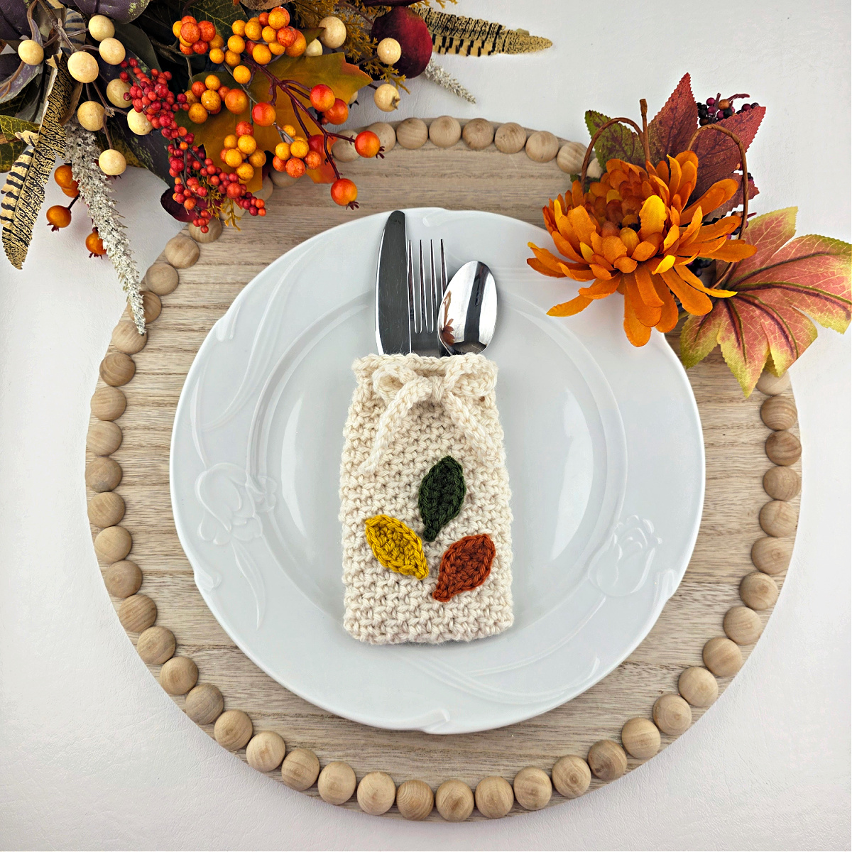 Orange, yellow, and green crochet leaves on a white cutlery holder with silverware in it on a white plate.