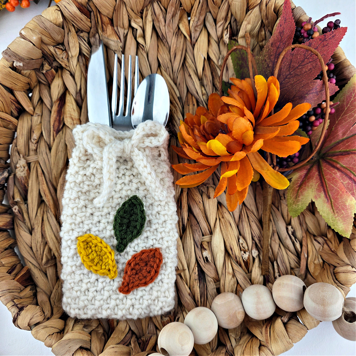 Orange, yellow, and green crochet leaves on a white cutlery holder with silverware in it on a brown background with wooden beads.