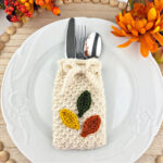 Orange, yellow, and green crochet leaves on a white cutlery holder with silverware in it on a white plate.