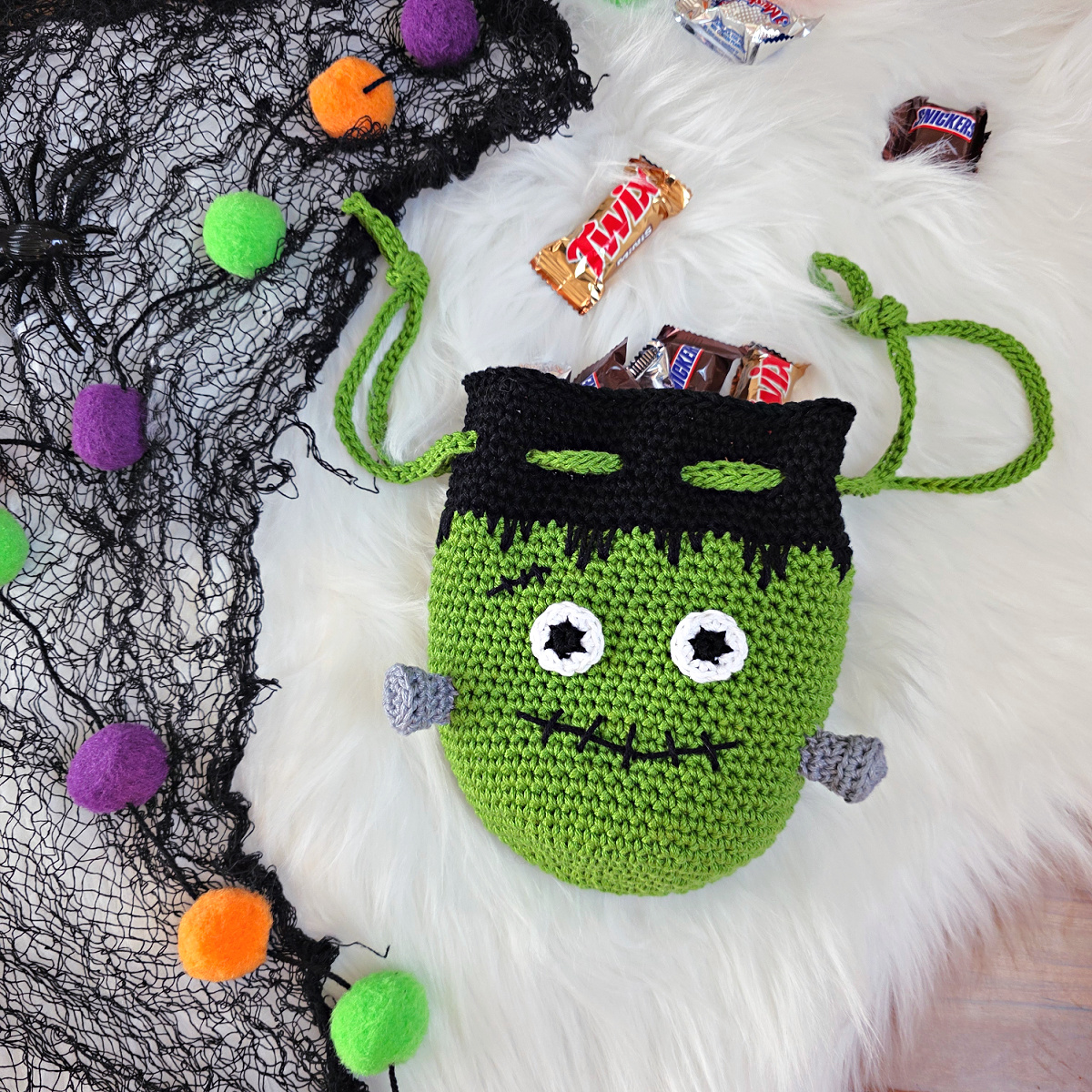 A frankenstein's monster crochet treat bag full of candy against a white faux fur and web background.
