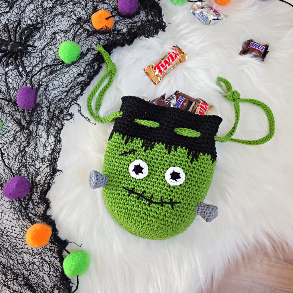 A frankensteins monster crochet treat bag full of candy against a white faux fur and web background.