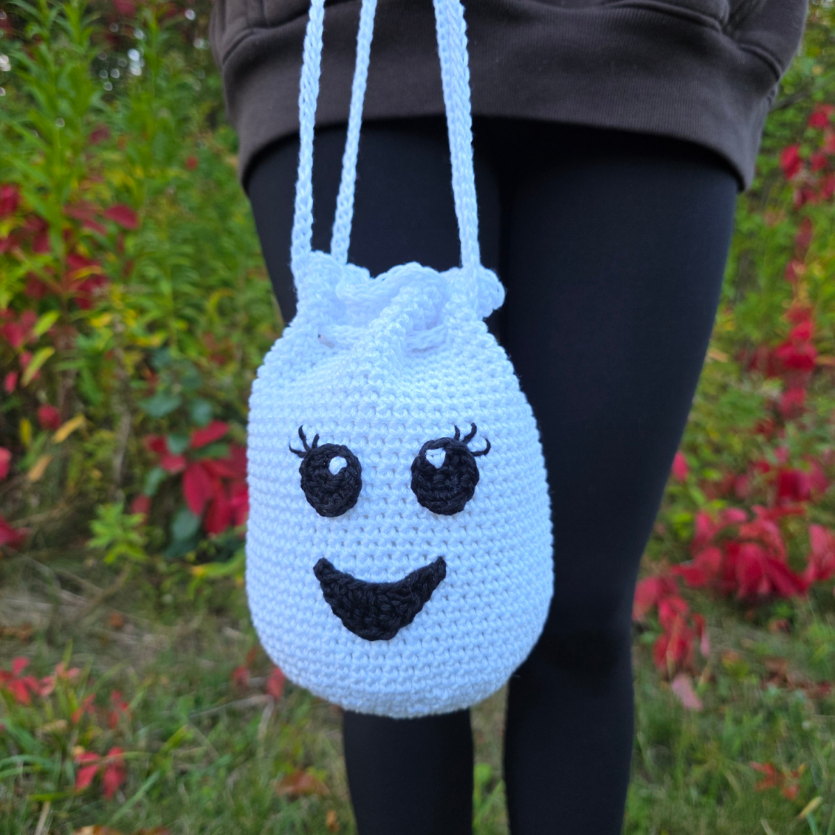 A person standing outside holding a ghost with a smiley face drawstring treat bag.