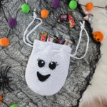 A crochet ghost treat bag full of candy against a white faux fur and web background.