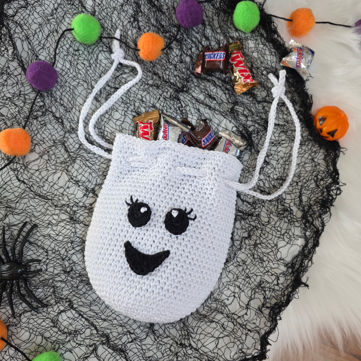 A crochet ghost treat bag full of candy against a white faux fur and web background.